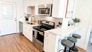 A modern kitchen with white cabinets, a stainless steel oven, a microwave, and a sink. There are three bar stools and a small potted plant on the counter.