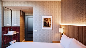 Habitación de hotel con una cama grande, un baño con lavamanos y espejo, un cuadro enmarcado en la pared y un cabecero de madera.