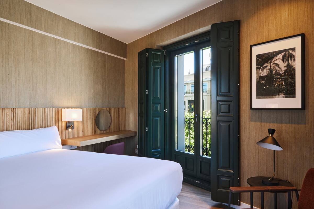 A hotel room with a bed, a desk with a lamp, a chair, and a door leading to an outdoor area.