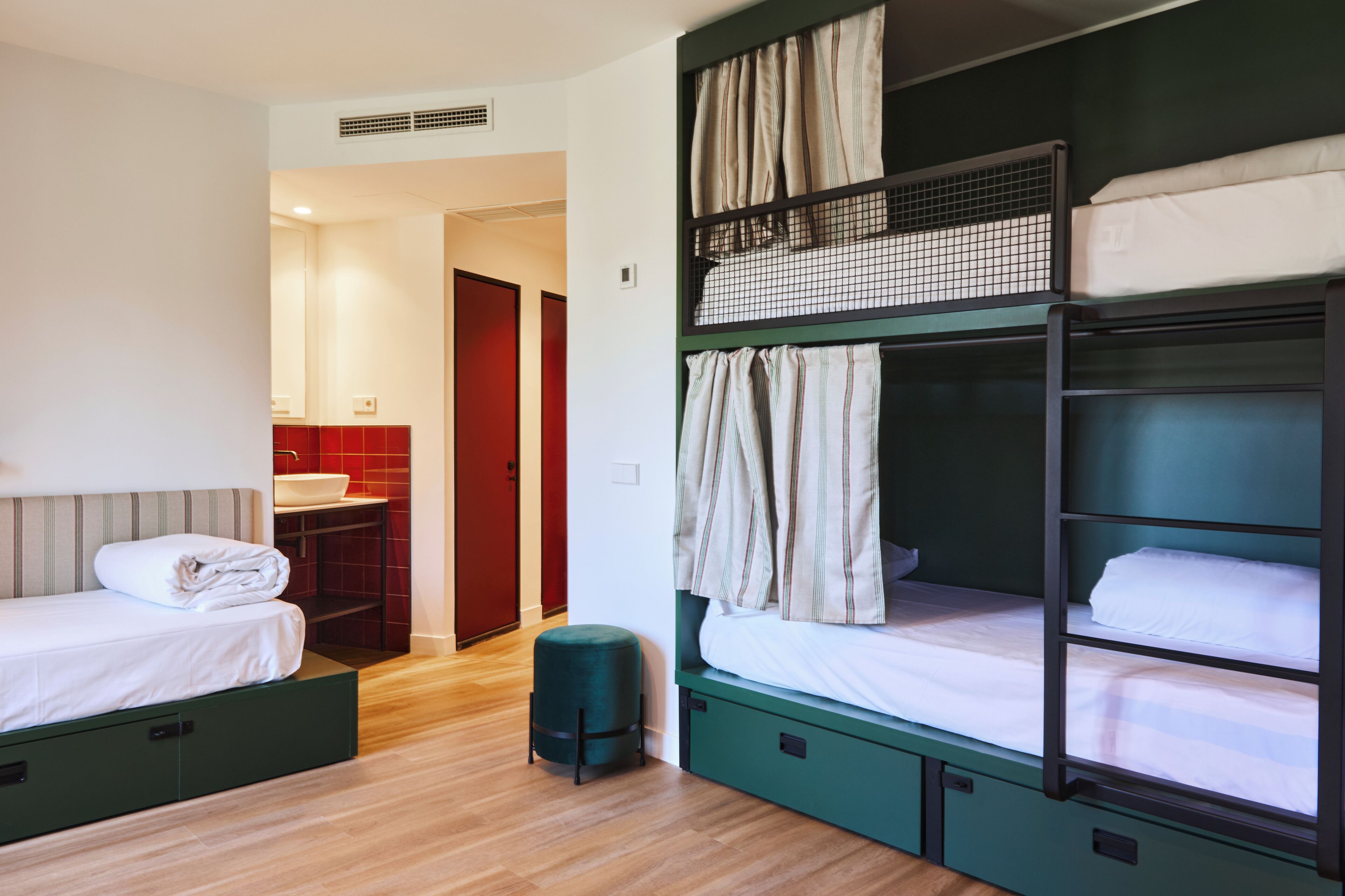 Una habitación tipo dormitorio con literas, un taburete pequeño y un baño con un lavabo rojo.