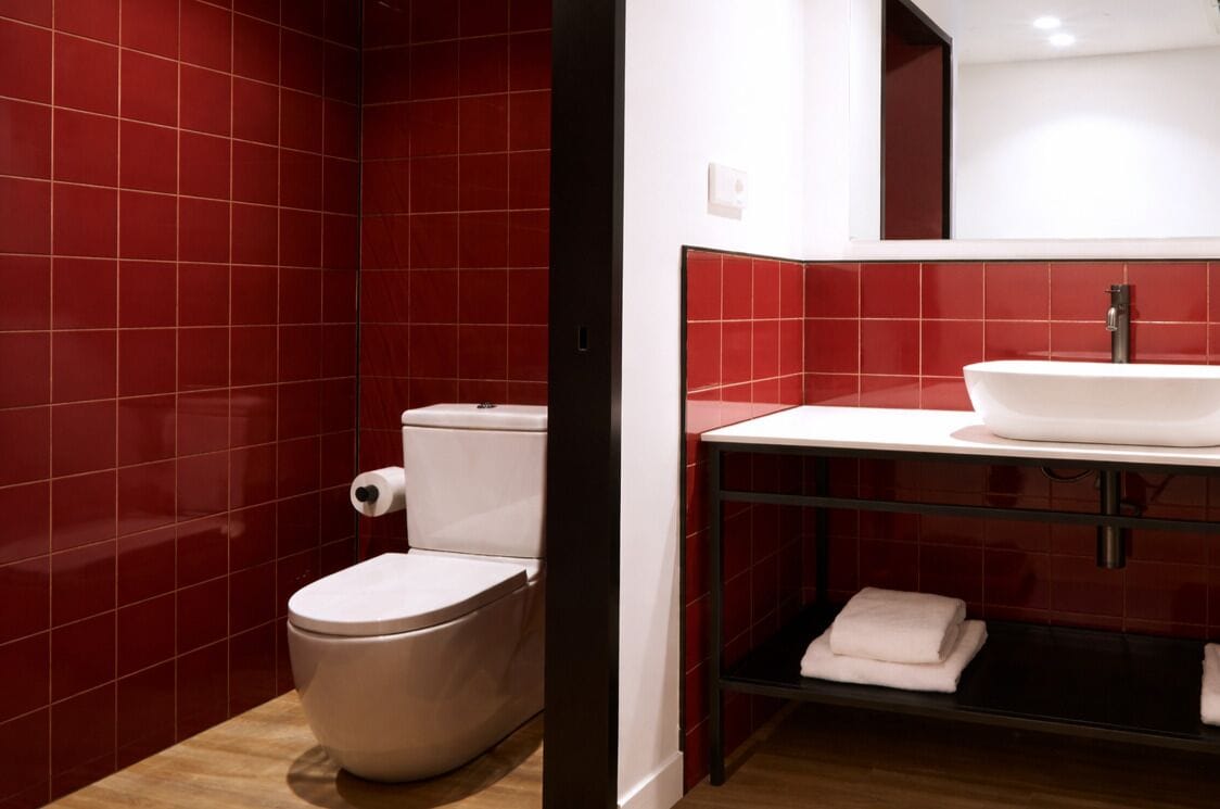 A bathroom with red tiles, a white toilet, a white sink, and a shelf with folded towels.