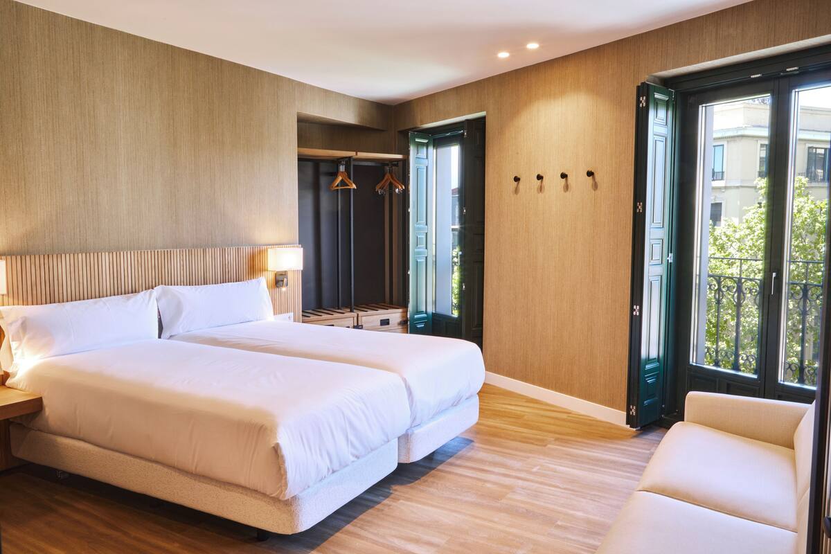 A hotel room with two beds, a wooden floor, a large window, and a view of the outside.