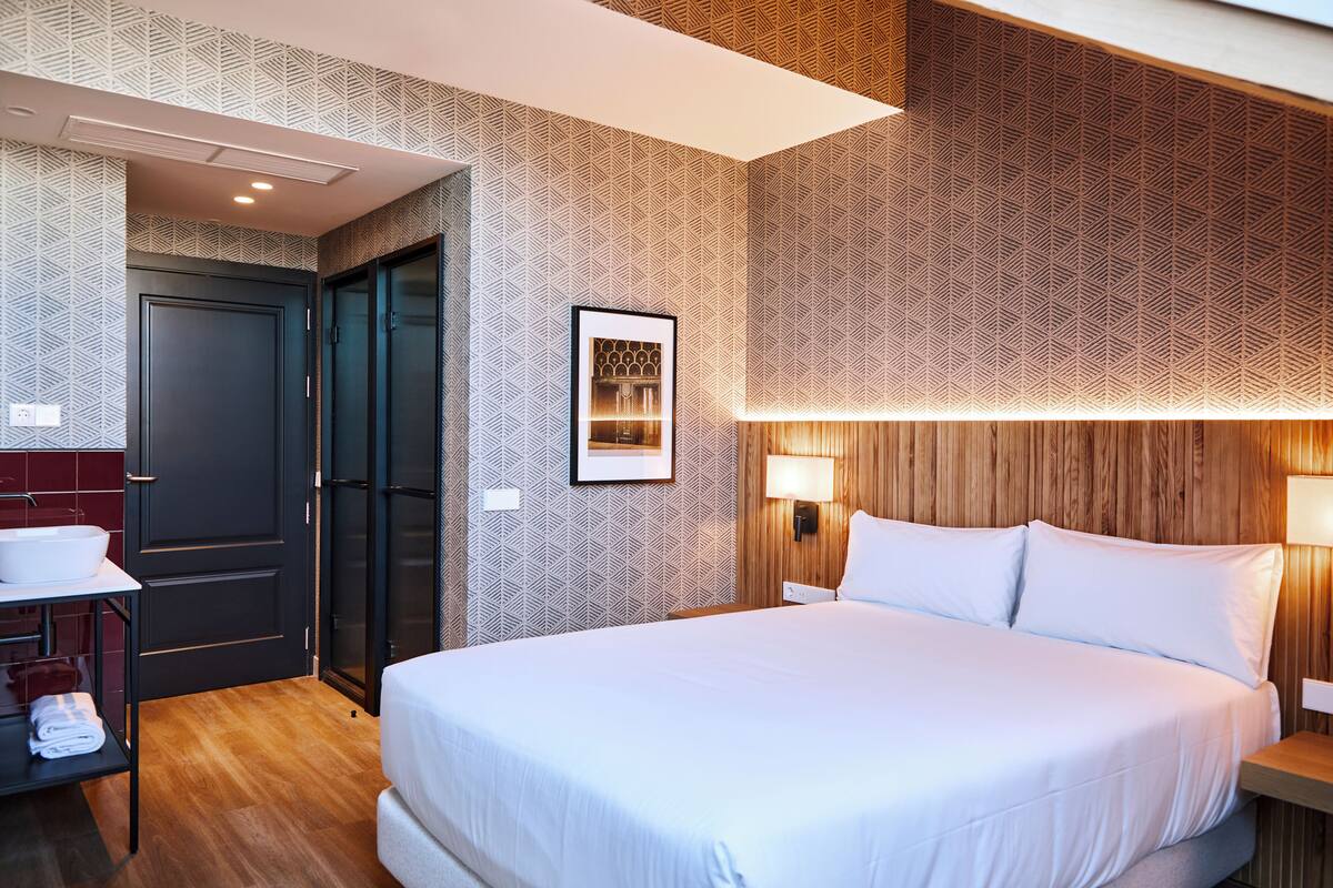 A hotel room with a large bed, a bedside table, a wall-mounted lamp, a framed picture, and a door leading to a bathroom.