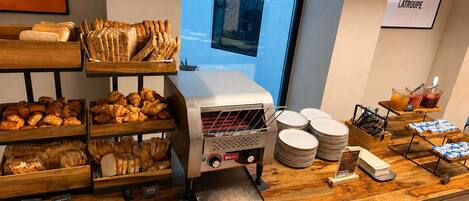 A breakfast buffet with various baked goods, a coffee machine, and a view of the city through a window.