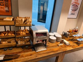 Ein Frühstücksbuffet mit verschiedenen Backwaren, einer Kaffeemaschine und Blick auf die Stadt durch ein Fenster.