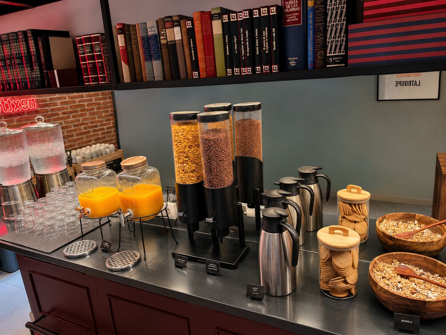 Una colazione a buffet con diversi distributori di cibo, una macchina per il caffè e una selezione di libri appesi al muro.