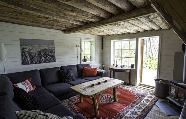 Ein gemütliches Wohnzimmer mit einem dunkelgrauen Sofa, einem Holztisch und einem roten Teppich mit Muster. Ein großes Fenster bietet Blick ins Grüne.