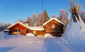 Eine Holzhütte mit verschneietem Dach und Schornstein, umgeben von Schnee und Bäumen.