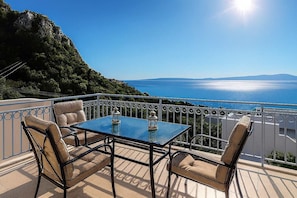A balcony with a table and chairs, overlooking a scenic view of the sea and mountains.