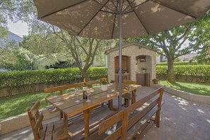Eine Außenpatio mit Holztisch und -bänken, einer Küchenzeile aus Stein und einem großen Sonnenschirm, der Schatten spendet.