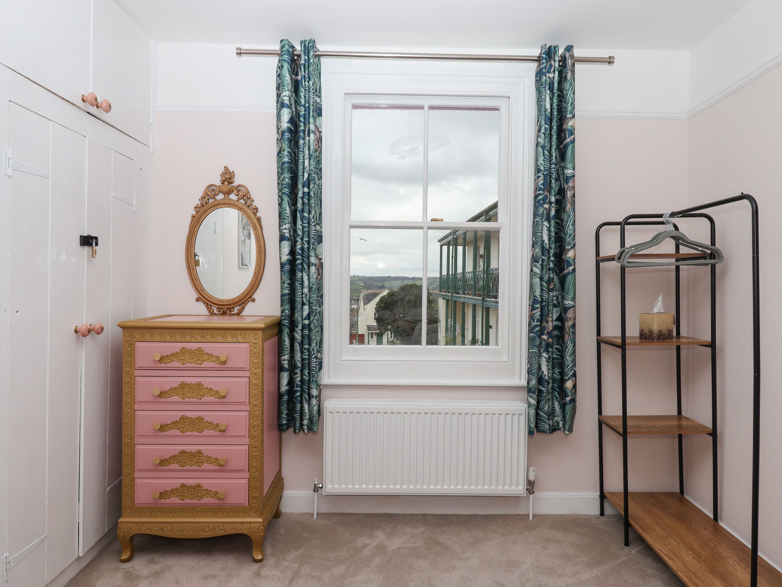 A room with a pink dresser, a mirror, a window with curtains, a radiator, and a metal shelf unit.