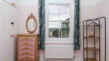 A room with a pink dresser, a mirror, a window with curtains, a radiator, and a metal shelf unit.