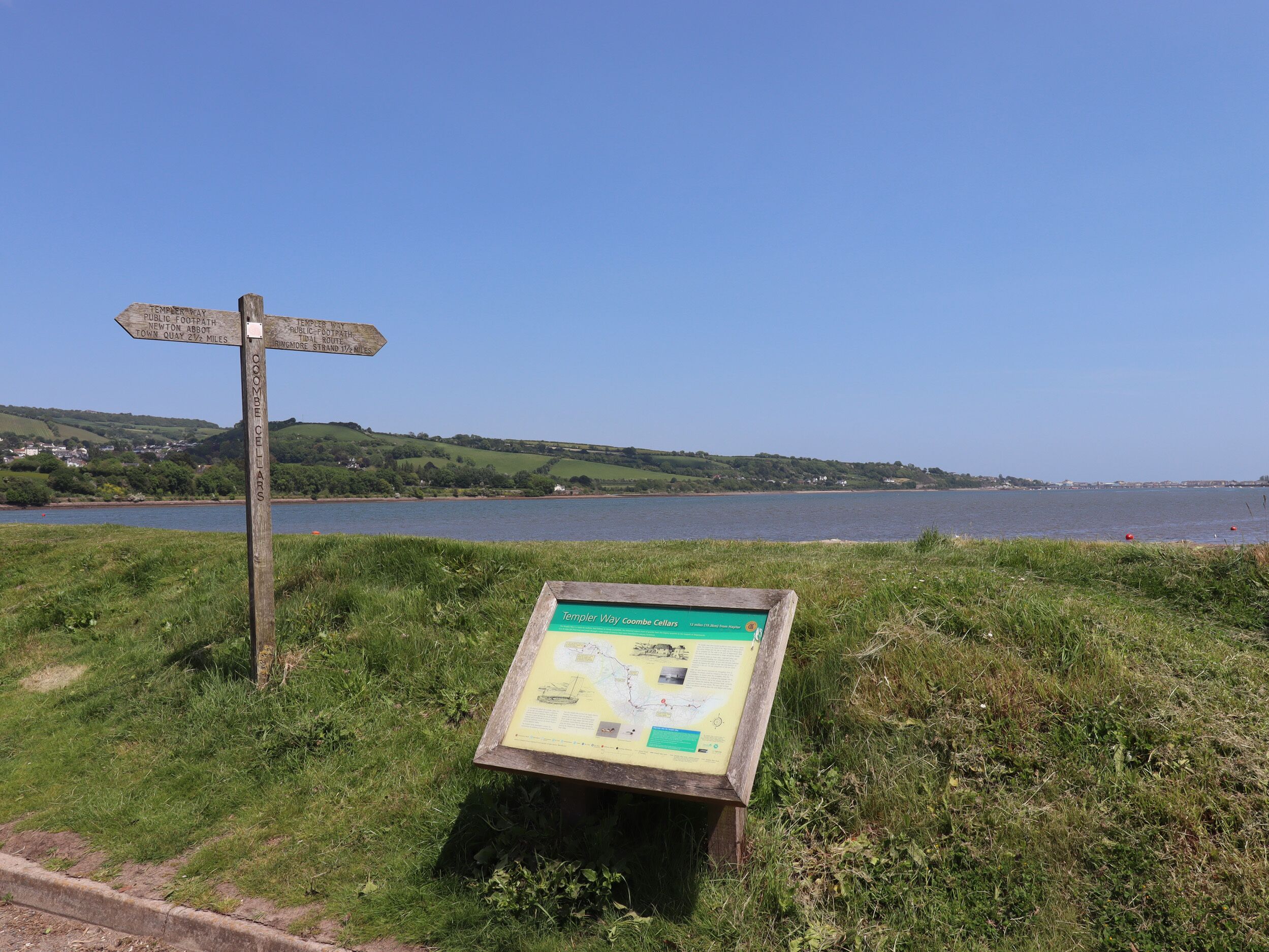 A wooden signpost with directional information and a map display board in a grassy area by a body of water.