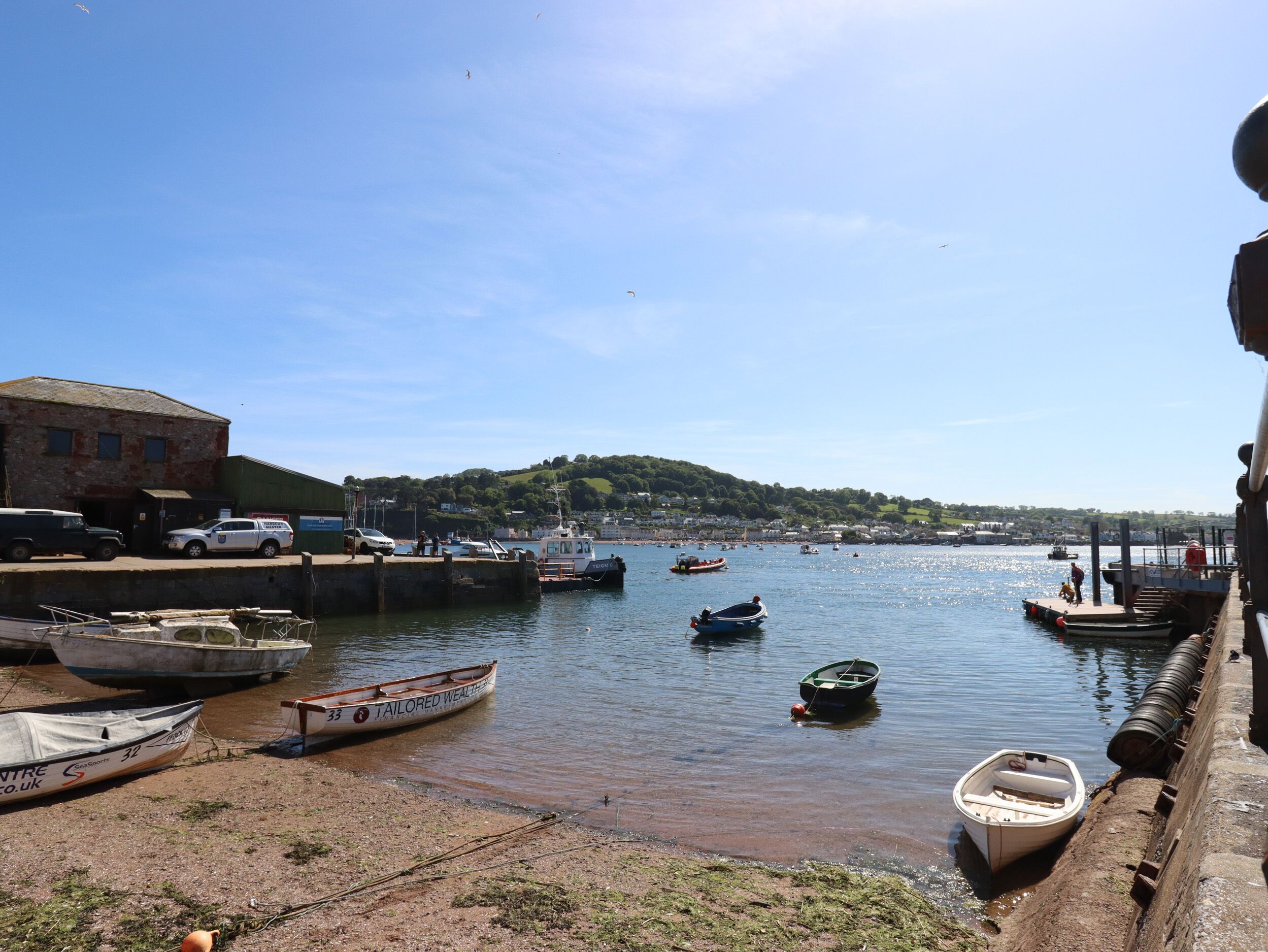 Un port avec des bateaux amarrés, un bâtiment sur la gauche et un paysage vallonné en arrière-plan.