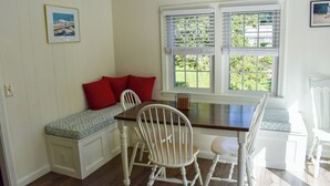 A dining area with a wooden table and white chairs, a bench with red cushions, and a window with blinds. - Renovated three bedroom home located in desirable Bayside Neighborhood! (Dennis)