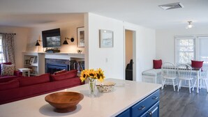 A modern kitchen with a white countertop, a wooden bowl, and a vase of sunflowers. There is a red sofa and a dining area with white chairs. - Renovated three bedroom home located in desirable Bayside Neighborhood! (Dennis)