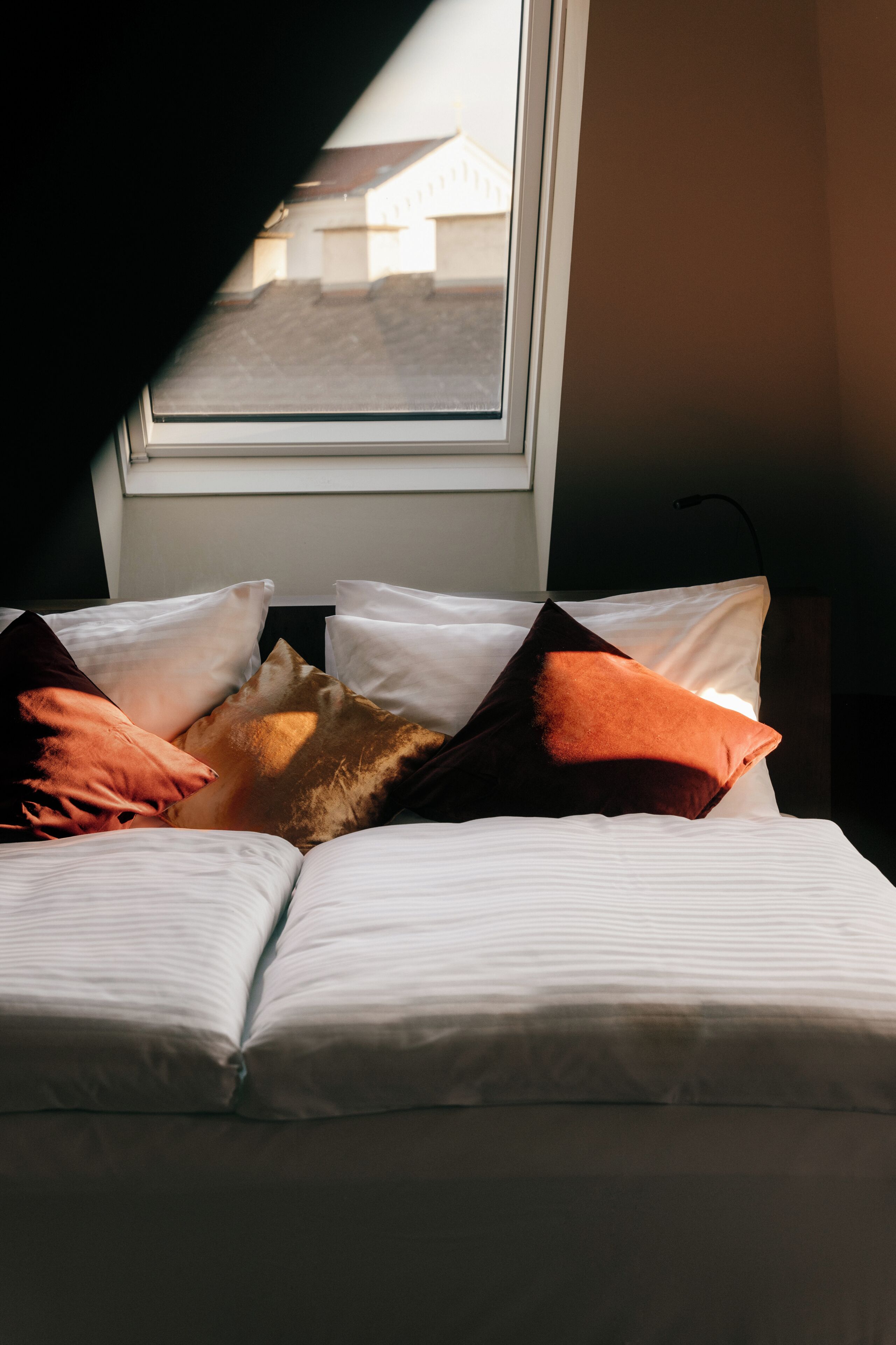 Un lit avec une literie blanche et plusieurs oreillers, dont certains d’une teinte brunâtre, placé sous une fenêtre donnant sur un bâtiment à l’extérieur.