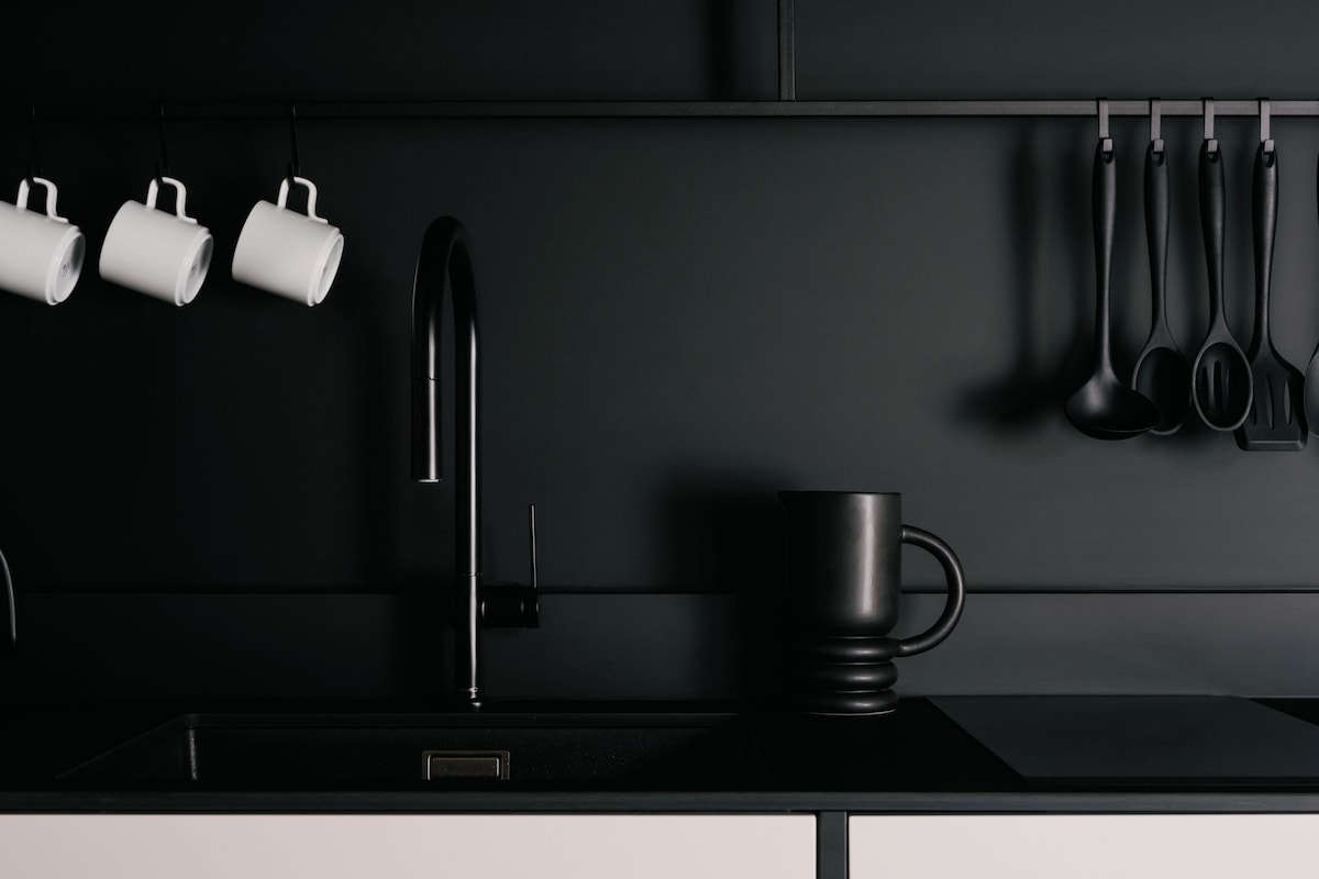 Une cuisine moderne avec un évier noir, un robinet noir et une tasse noire. Trois tasses blanches sont accrochées au mur, et plusieurs ustensiles de cuisine sont suspendus au-dessus de l’évier.
