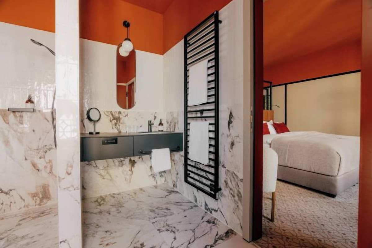 Une salle de bain moderne avec un sol en marbre, un porte-serviettes noir et un lit recouvert d’une courtepointe blanche dans la pièce adjacente.