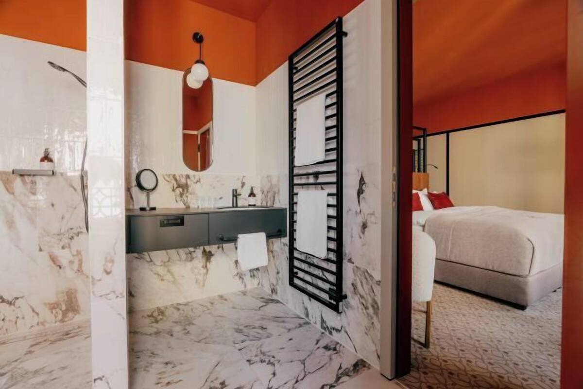 Een moderne badkamer met een marmeren vloer, een zwarte handdoekhouder, en in de aangrenzende kamer een bed met een witte dekbedovertrek.