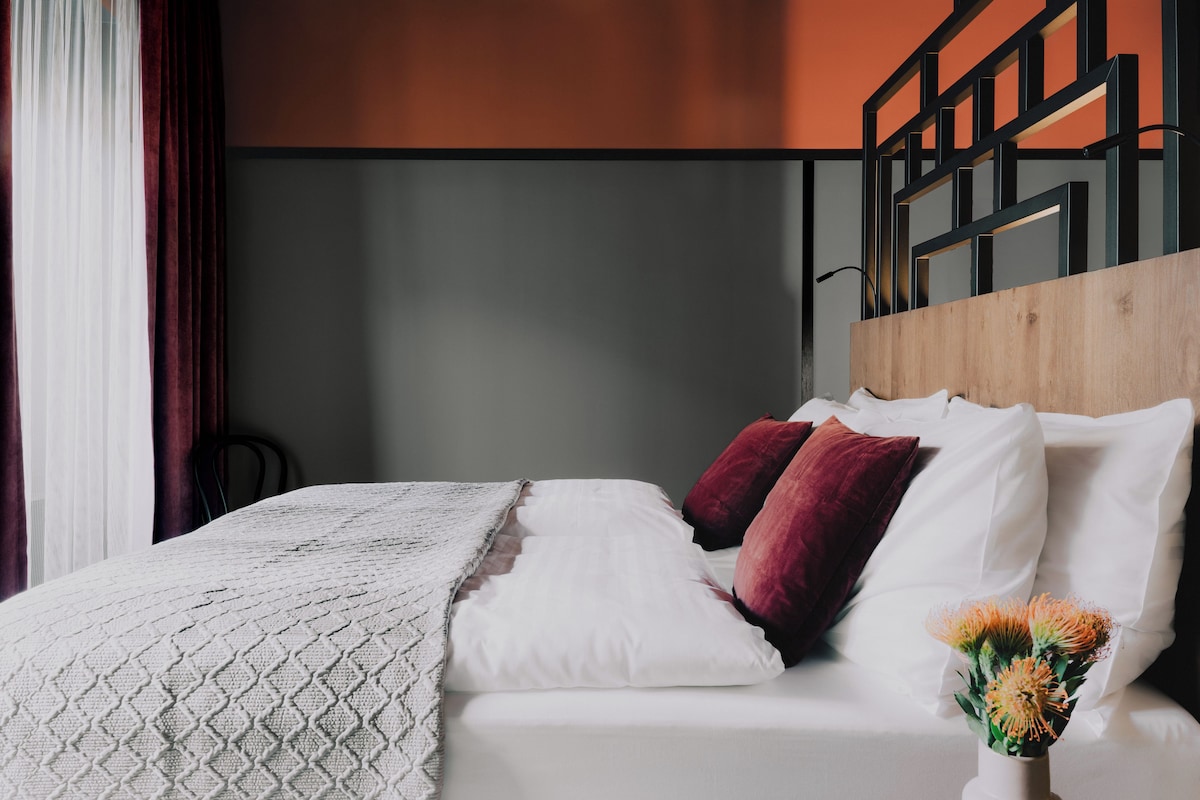 Un lit bien fait, avec des oreillers blancs et rouges, une couverture blanche et un vase rempli de fleurs, le tout posé sur une tête de lit en bois.