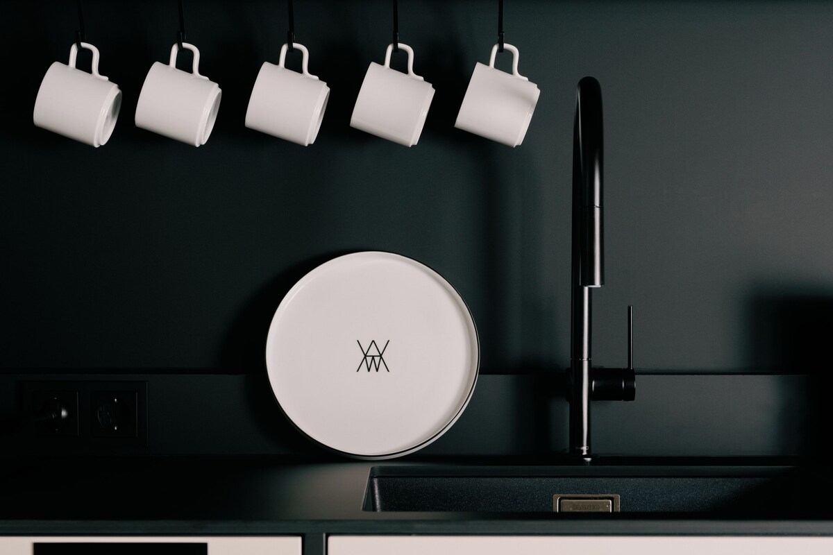 Une cuisine au design moderne, avec un appareil électroménager blanc et de forme circulaire orné d’un logo, un robinet noir et cinq tasses blanches suspendues à un porte-tasses noir.