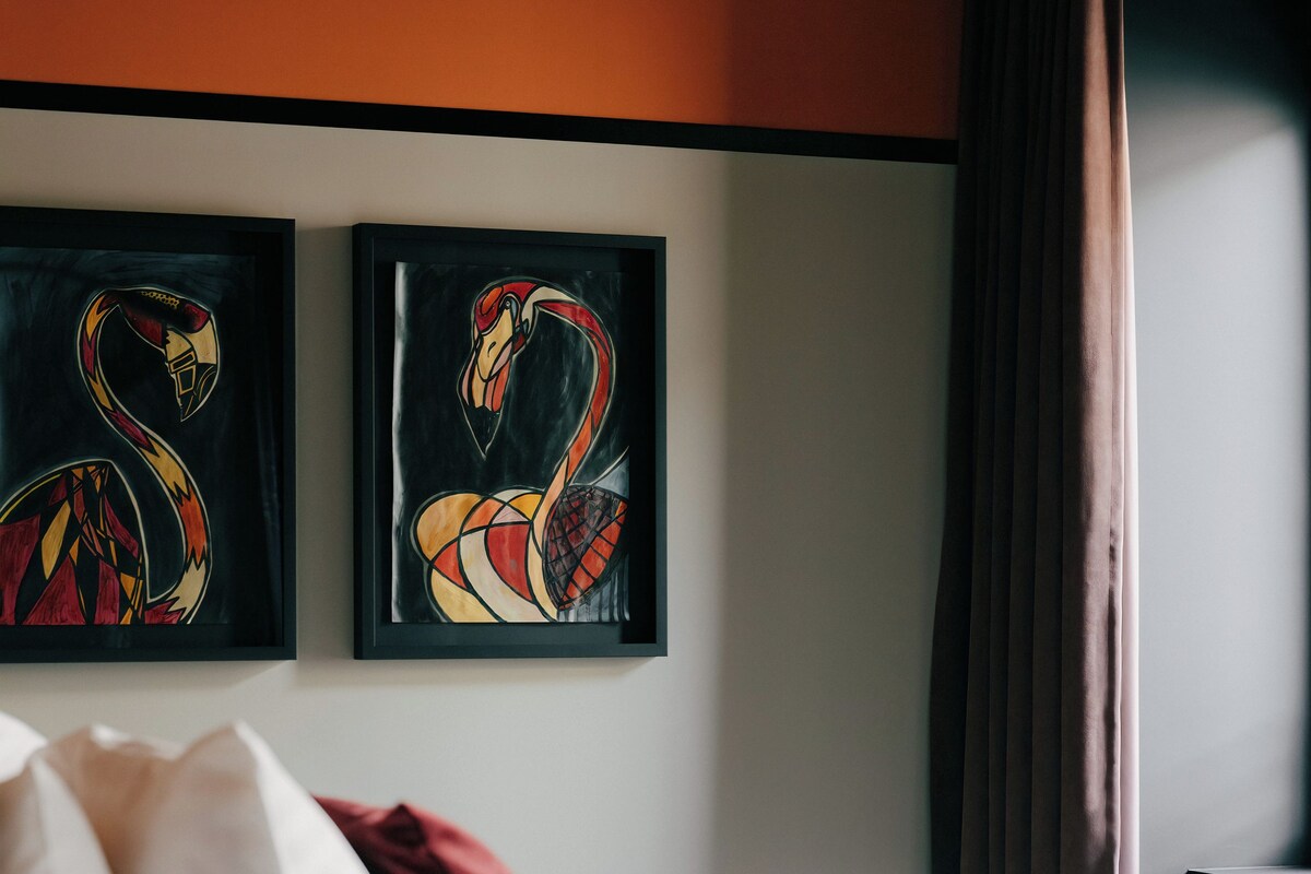 Hotelkamer met een bed, witte beddengoed, een rood sierkussen en twee flamingo’s aan de muur bevestigde schilderijen.