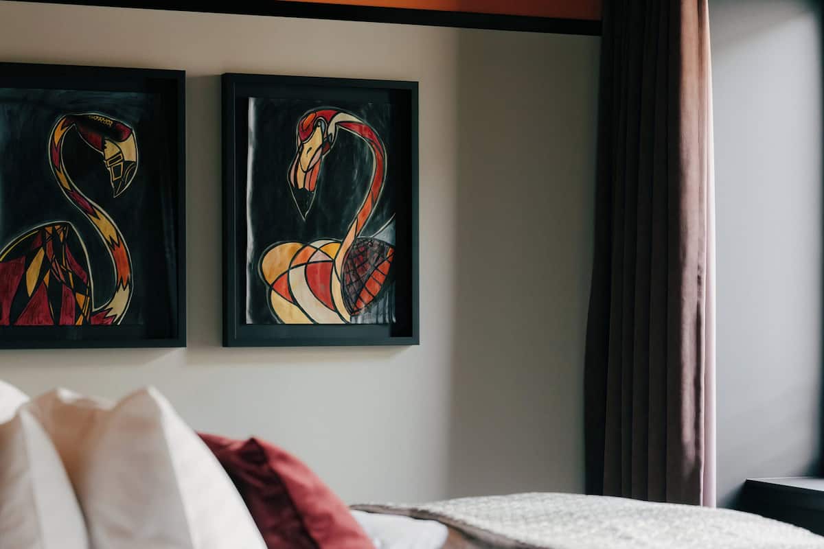 Une chambre d’hôtel avec un lit, du linge de lit blanc, un coussin rouge et deux tableaux représentant des flamants roses, accrochés au mur.