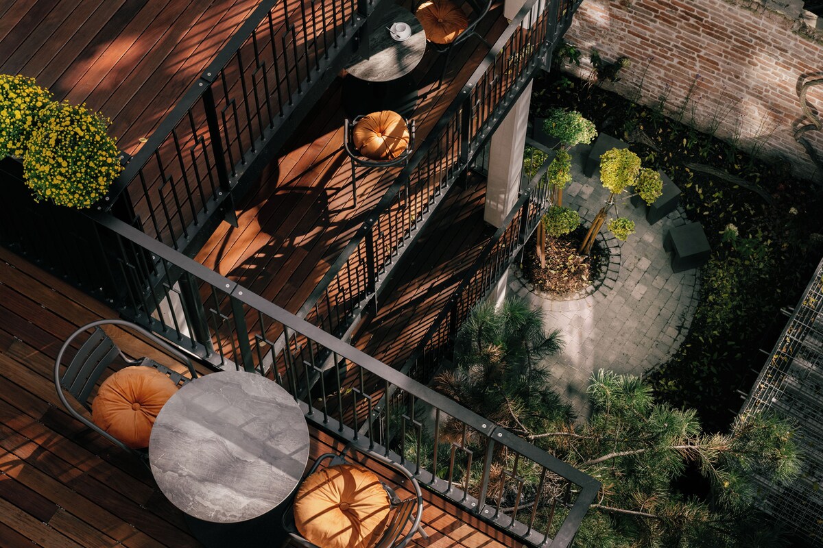 Een terras buiten met meerdere niveaus, houten vloer, metalen reling en diverse zithoekjes, waaronder ronde tafels met stoelen.