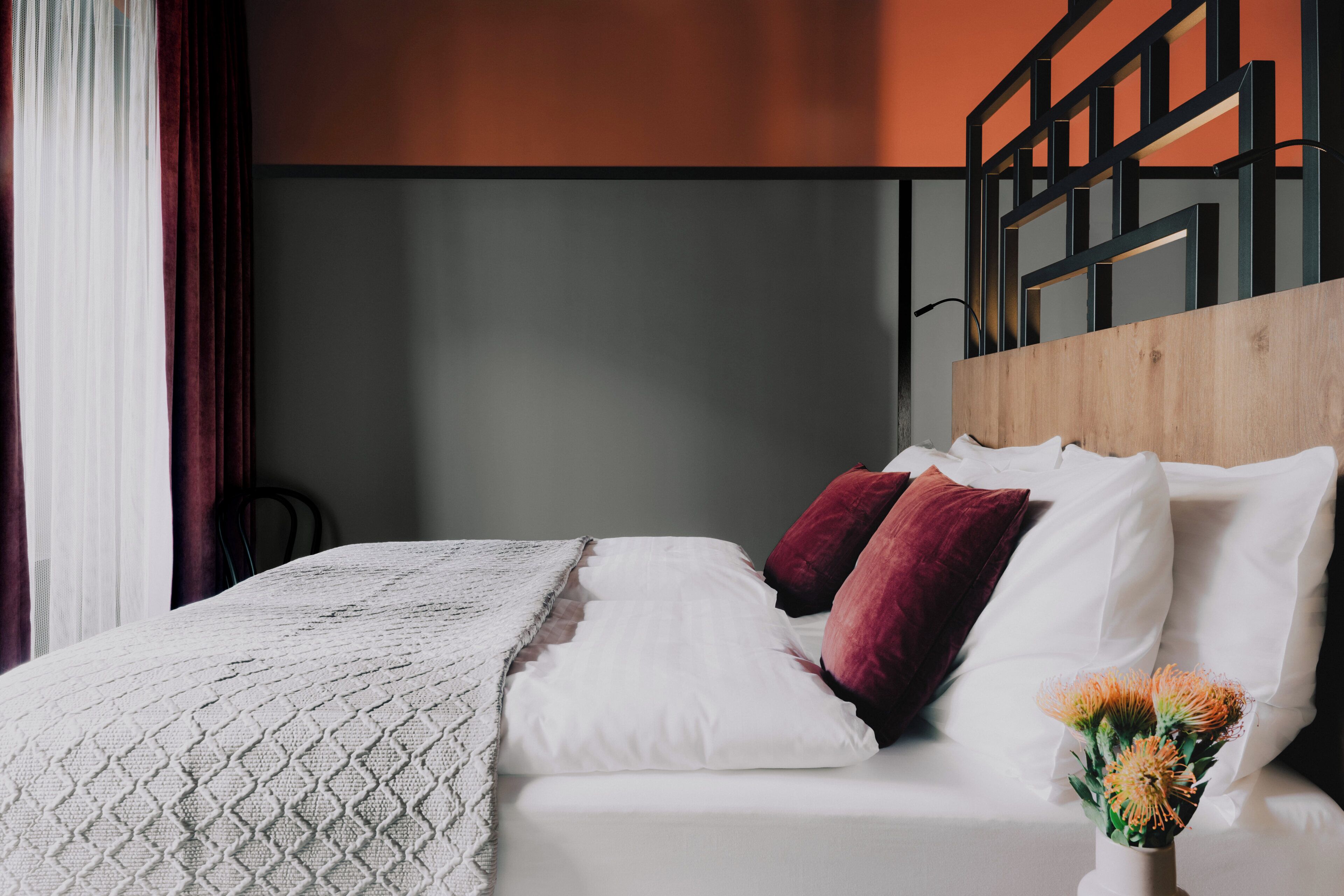 Un lit bien fait, avec des oreillers blancs et rouges, une couverture blanche et un vase rempli de fleurs, le tout posé sur une tête de lit en bois.