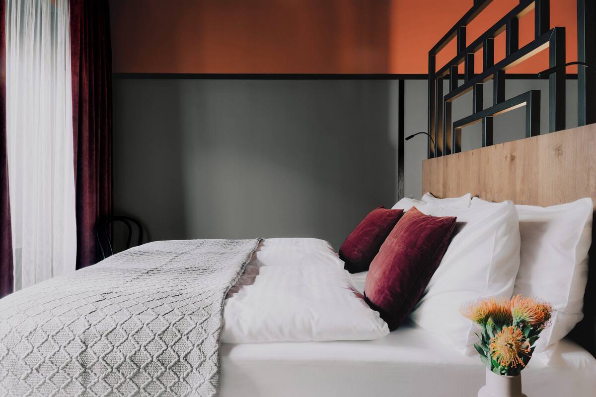 Een net opgemaakt bed met witte en rode kussens, een witte deken en een vaas met bloemen op een houten hoofdbord.