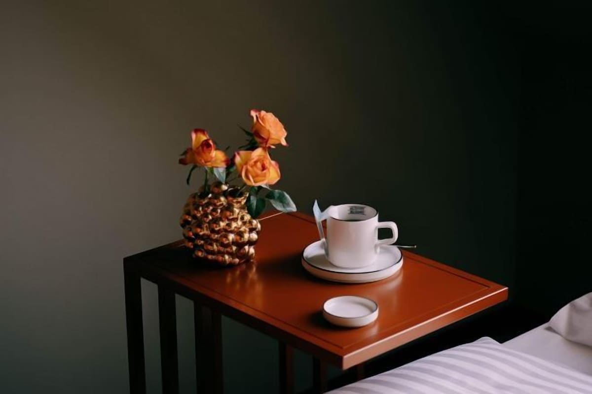 Une table de chevet en bois sur laquelle se trouvent une tasse et une soucoupe blanches, un ananas et un vase rempli de fleurs.