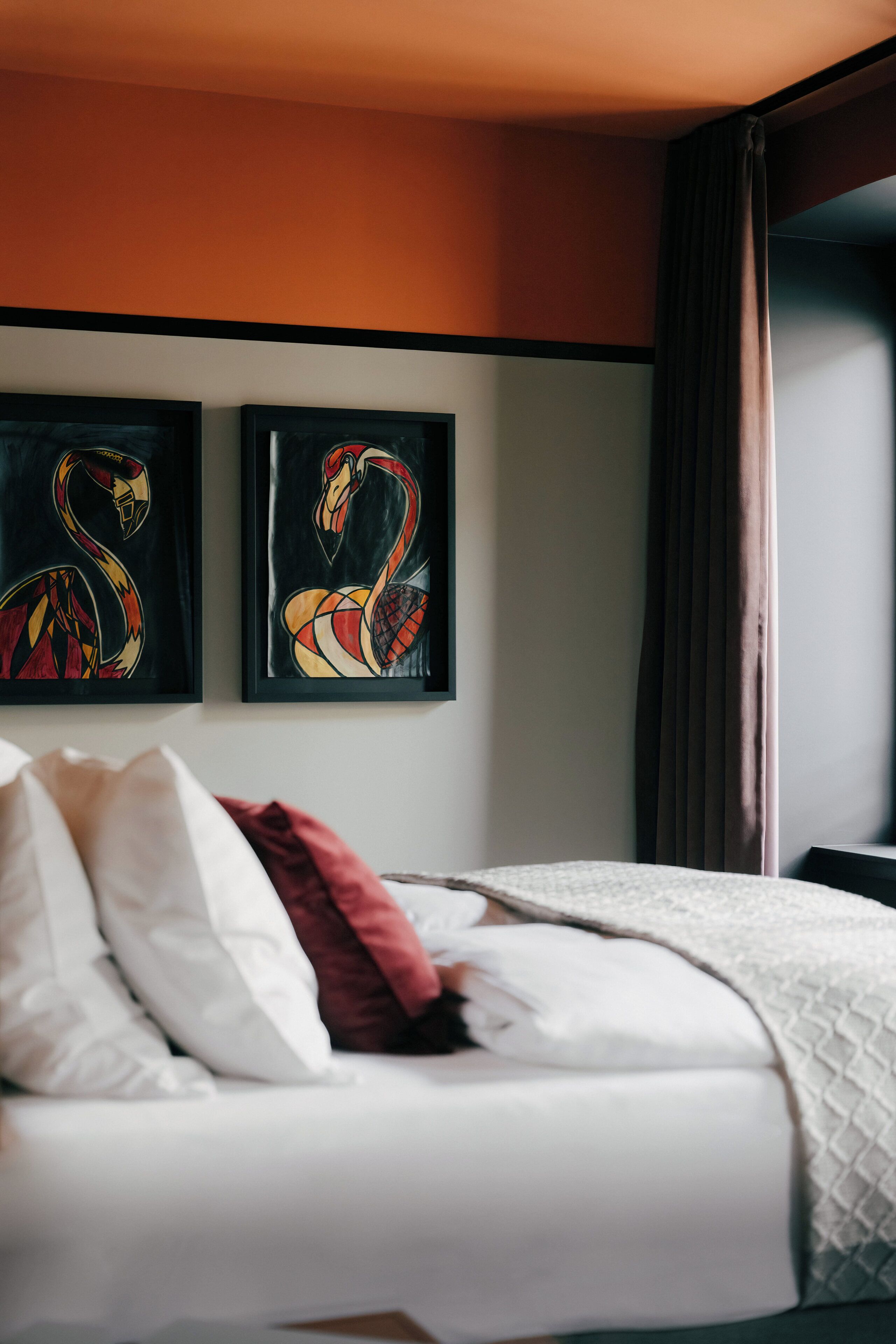 Une chambre d’hôtel avec un lit, du linge de lit blanc, un coussin rouge et deux tableaux représentant des flamants roses, accrochés au mur.