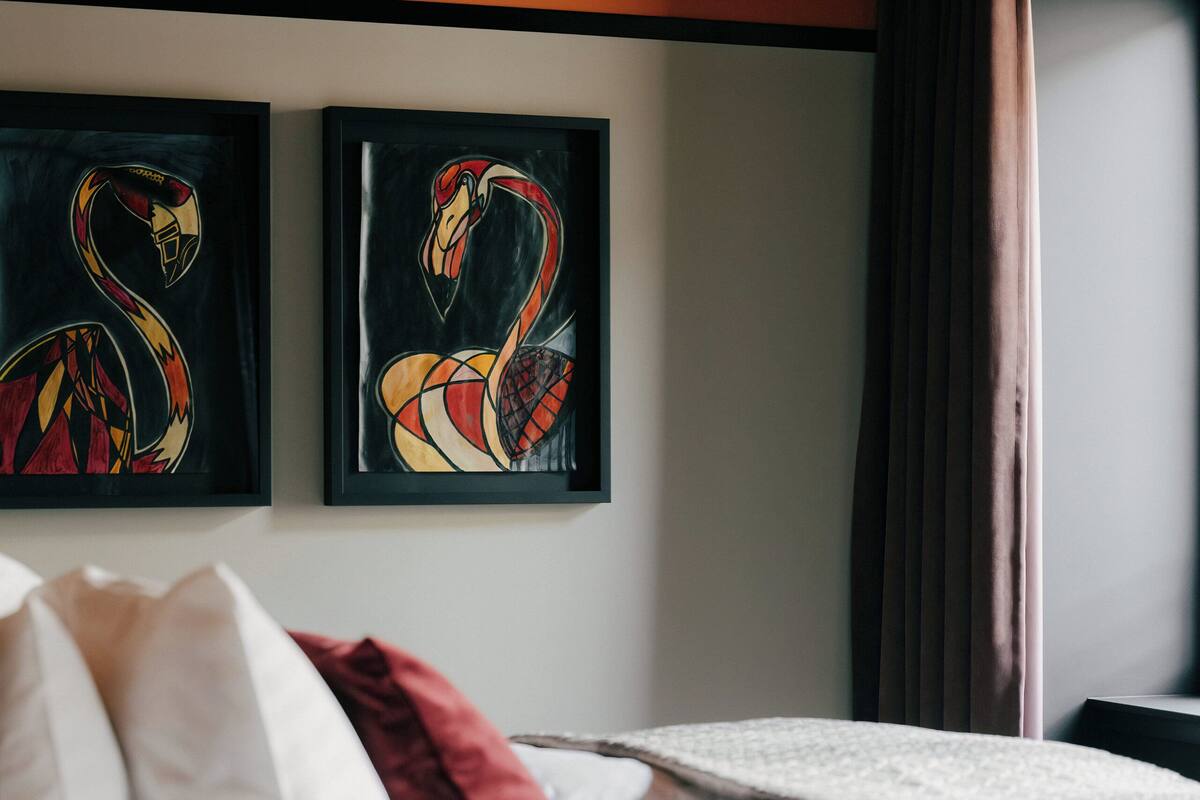Hotelkamer met een bed, witte beddengoed, een rood sierkussen en twee flamingo’s aan de muur bevestigde schilderijen.