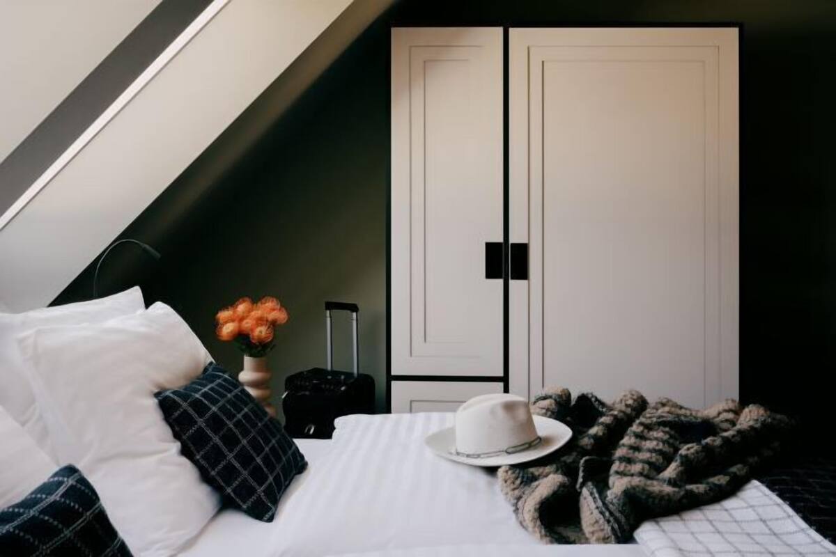 Een slaapkamer met een bed, kussens, een koffer, een hoed, een deken en een kledingkast.
