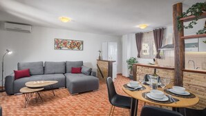 Una sala de estar moderna con un sofá gris, una mesa de comedor para cuatro personas y una zona de cocina con un salpicadero de ladrillo.