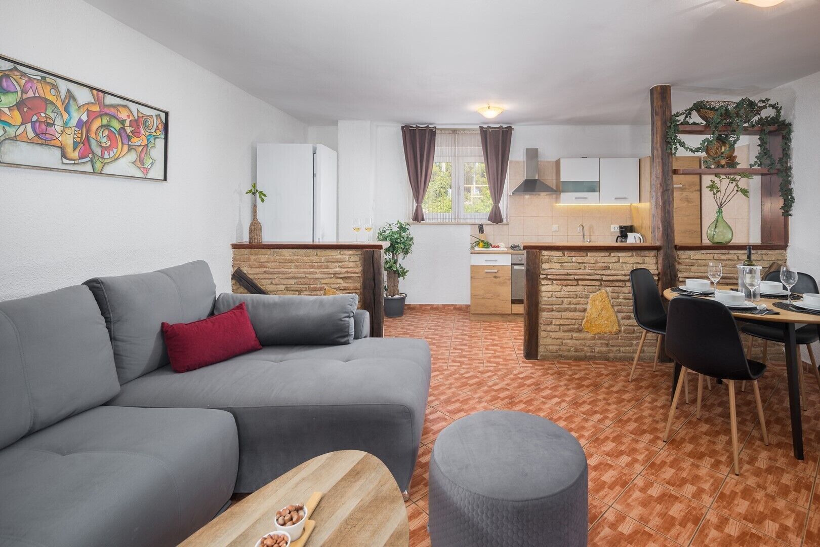 A modern living room with a grey sofa, a small round ottoman, and a dining area with a wooden table and chairs.