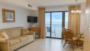 A hotel room with a sofa, a dining table with chairs, a TV, and a balcony with curtains. - Oceanfront Efficiency direct ocean views pool, gym & near all (Ocean City)