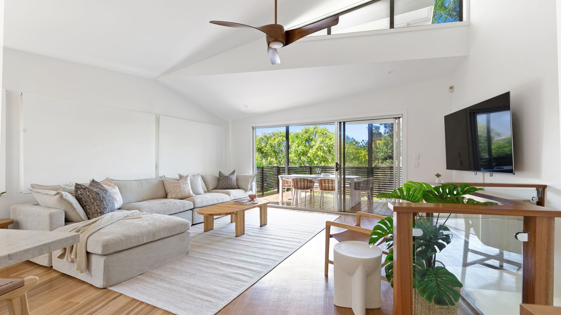 A modern living room with a sofa, coffee table, and dining area. There is a TV mounted on the wall and a ceiling fan.