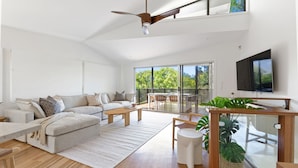Un salon moderne avec un canapé, une table basse et un coin repas. Un téléviseur est fixé au mur et un ventilateur de plafond est installé.