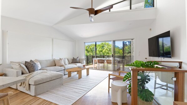 A modern living room with a sofa, coffee table, and dining area. There is a TV mounted on the wall and a ceiling fan.