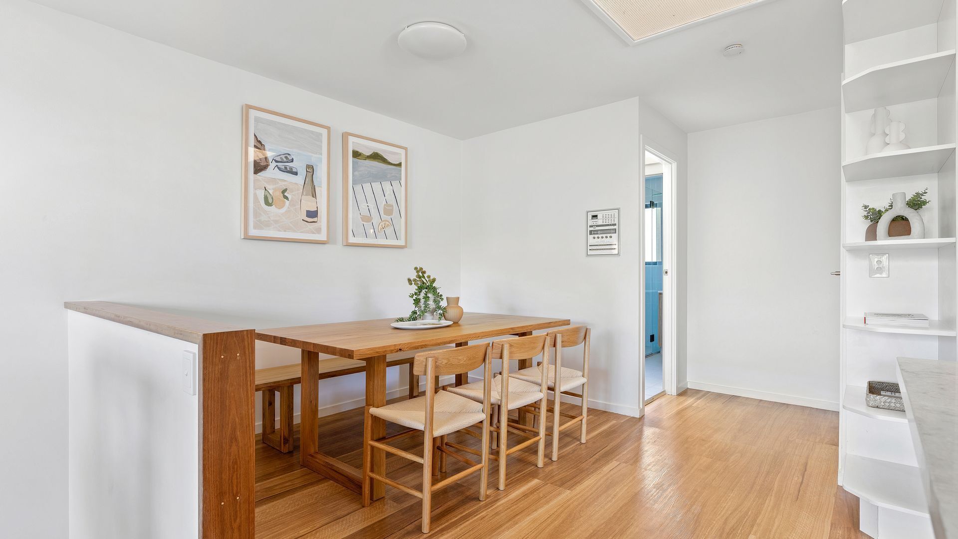 A dining area with a wooden table and chairs, a small plant on the table, and two framed pictures on the wall.