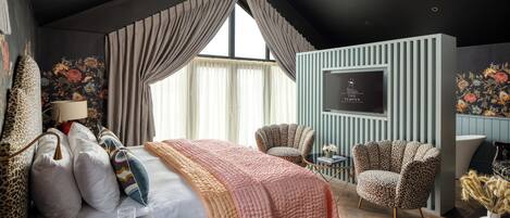 Ein Hotelzimmer mit einem großen Bett, zwei Sesseln in Leopardenmuster, einem Fernseher und einem Schreibtisch mit Lampe und Büchern.