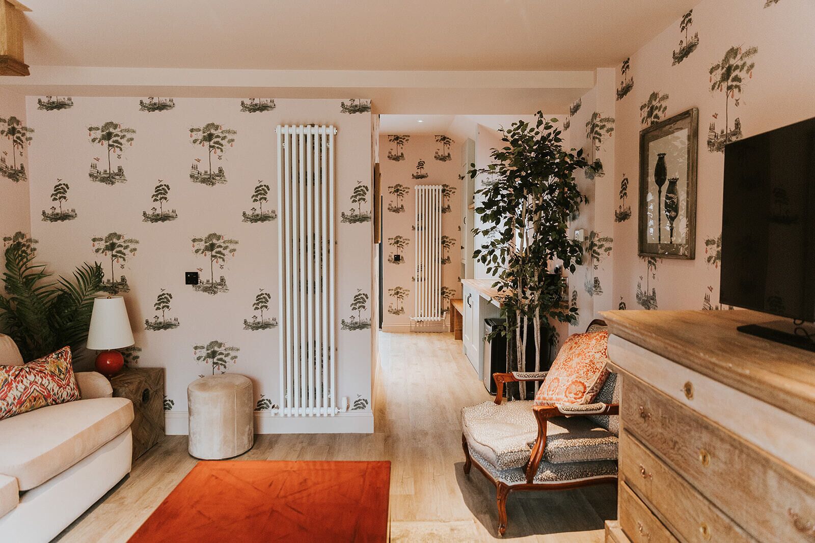 Un salon avec un canapé, un fauteuil et un téléviseur posé sur un meuble en bois. On y trouve également un radiateur, une plante et une lampe sur une table d’appoint.