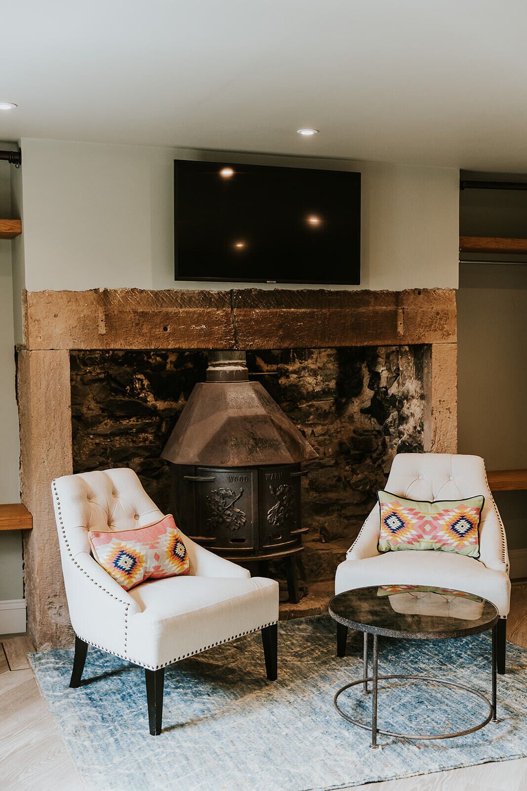 A cozy room with a stone fireplace, two upholstered chairs, a small round table, and a flat-screen TV mounted above the fireplace.