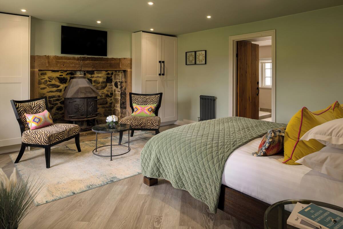 Un dormitorio con chimenea de piedra, dos sillones tapizados, una cama con colcha acolchada y una mesita con un florero.