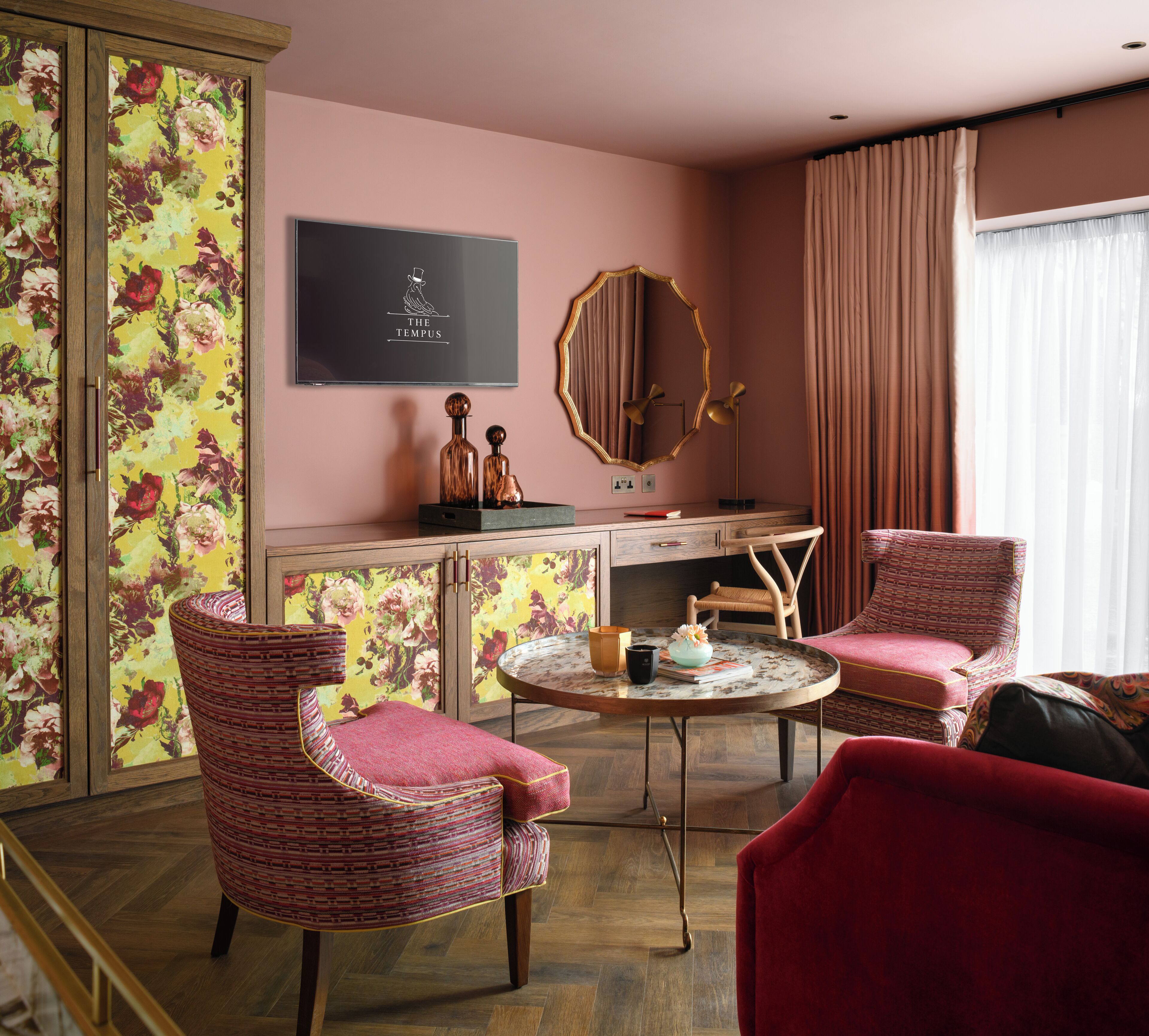 Una stanza con parete rosa, carta da parati floreale, un tavolo rotondo con sedie, uno specchio e una TV a schermo piatto che mostra 'The Tempus'.