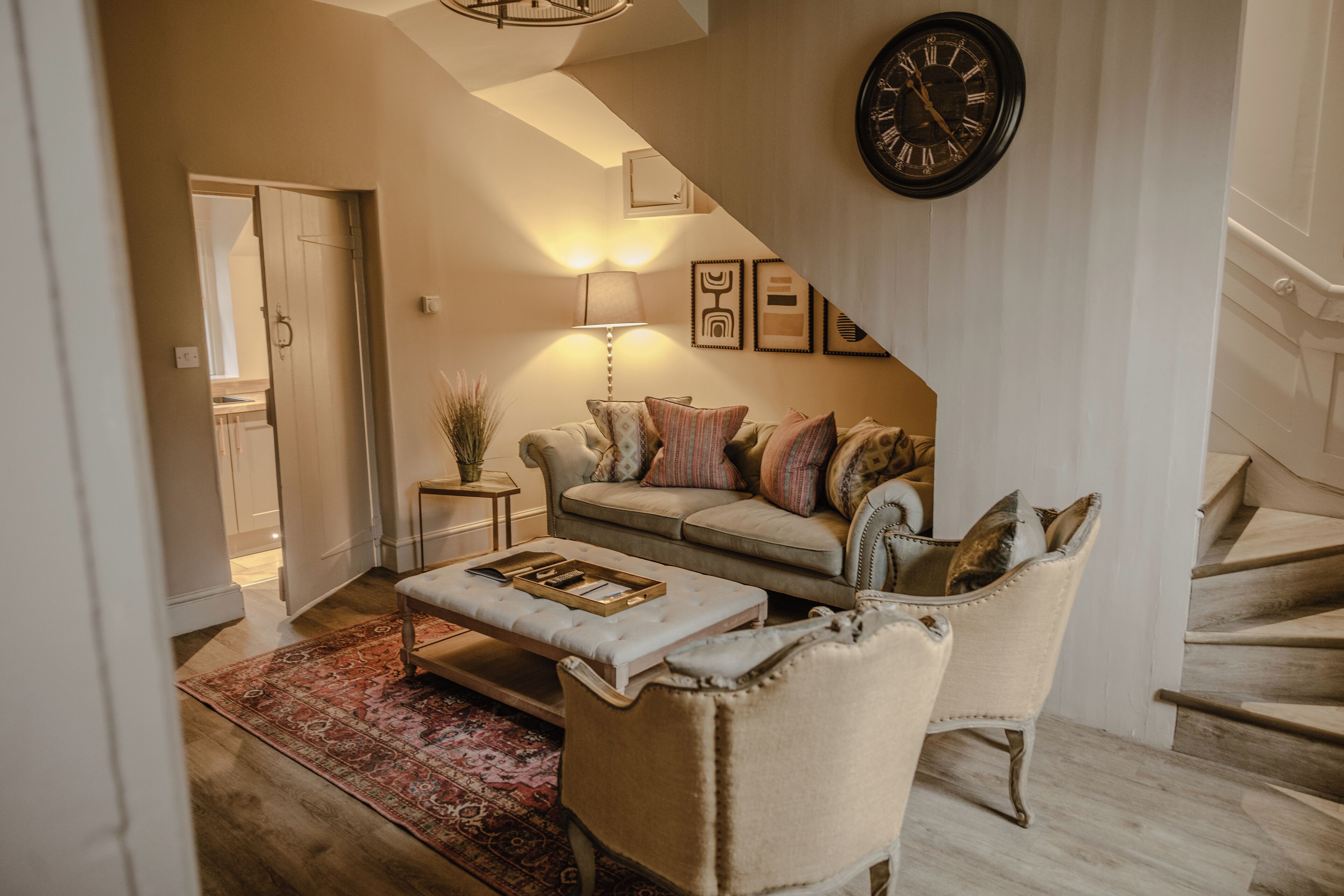 Una sala de estar con sofá, sillón y reposapiés, una escalera y un reloj de pared.