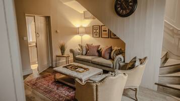 Una sala de estar con sofá, sillón y reposapiés, una escalera y un reloj de pared.