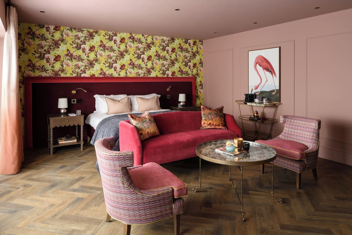 Una habitación de hotel con una cama grande, un sofá rojo, dos sillones, una mesita redonda y una pared con papel tapiz floral.
